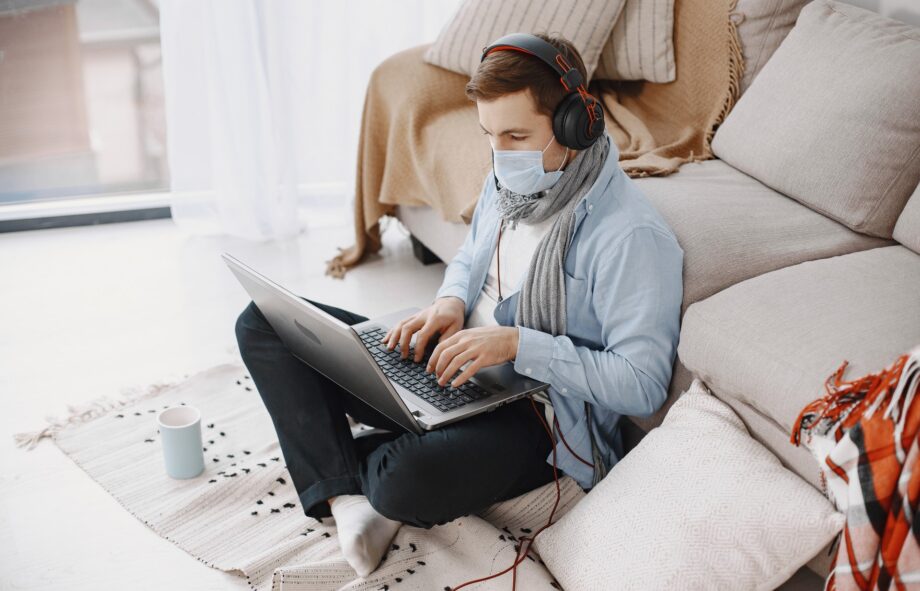 Male in a medical mask. Man sitting in living room at home. Guy enjoying studying on quarantine.